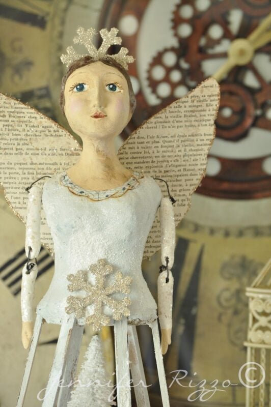 A handcrafted angel figurine inspired by Angel Santos Cage Dolls stands with paper wings before a blurred clock background. The artist's signature, Jennifer Rizzo, is visible on the image.