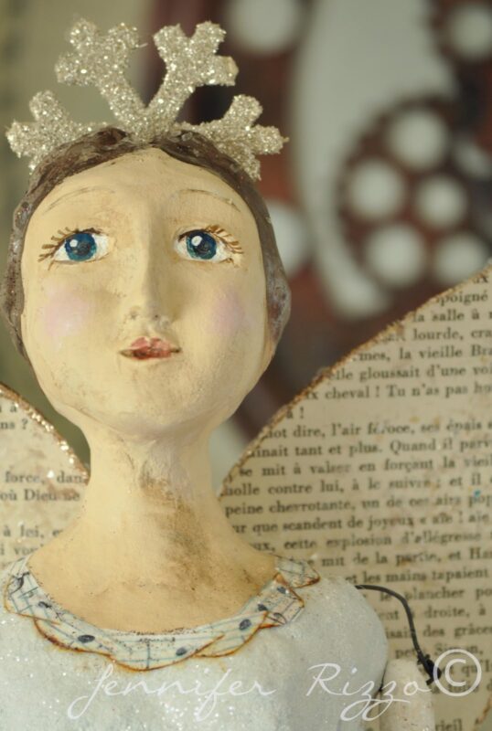A handmade angel figurine inspired by Angel Santos Cage Dolls, with a glittery crown and paper wings, featuring blue eyes and a painted face, stands in front of a blurred background.