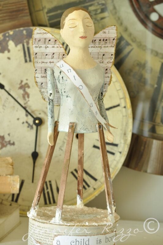 A handmade Angel Santos Cage Doll figurine with musical sheet wings stands on long legs in front of vintage clock faces, capturing timeless charm and artistry.