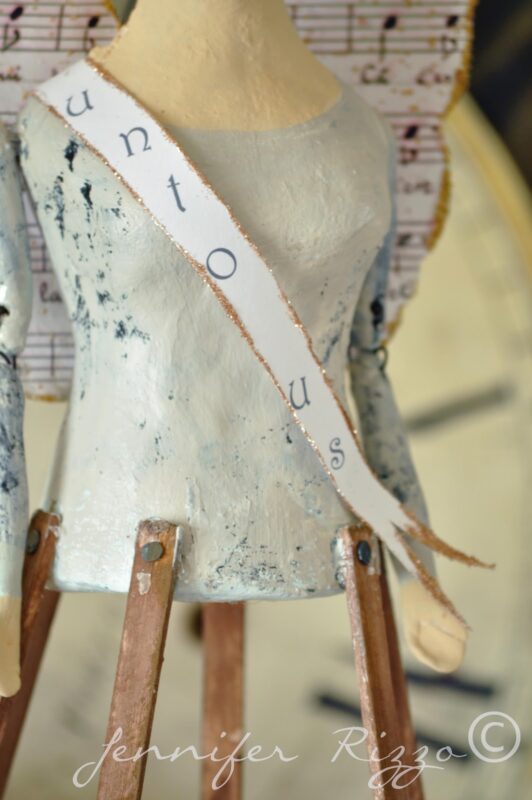 Close-up of a handmade Angel Santos Cage Doll figure with a white sash reading "untous," musical note wings, and wooden peg legs. "Jennifer Rizzo" is watermarked at the bottom. Perfect alongside handmade earrings.