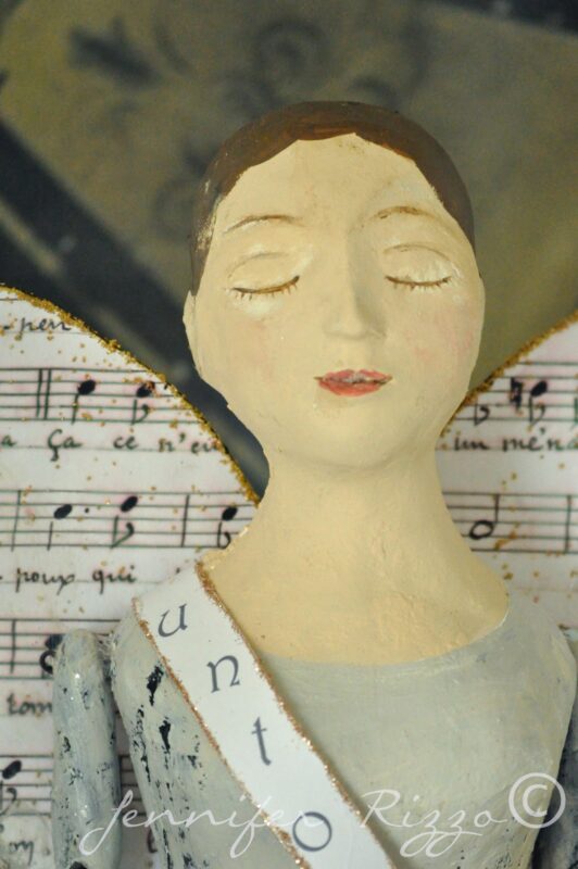A painted angel figurine with closed eyes, inspired by Angel Santos Cage Dolls, wears a sash with partial text and wings decorated with sheet music notes—perfect for pairing with handmade earrings.