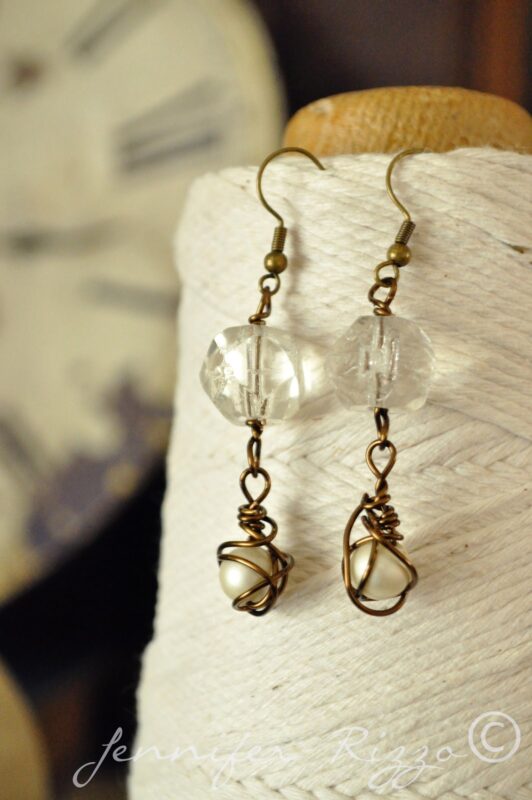 A pair of Handmade Earrings inspired by Angel Santos Cage Dolls, featuring clear beads and pearl-like stones wrapped in bronze wire, beautifully displayed on a spool of white string.