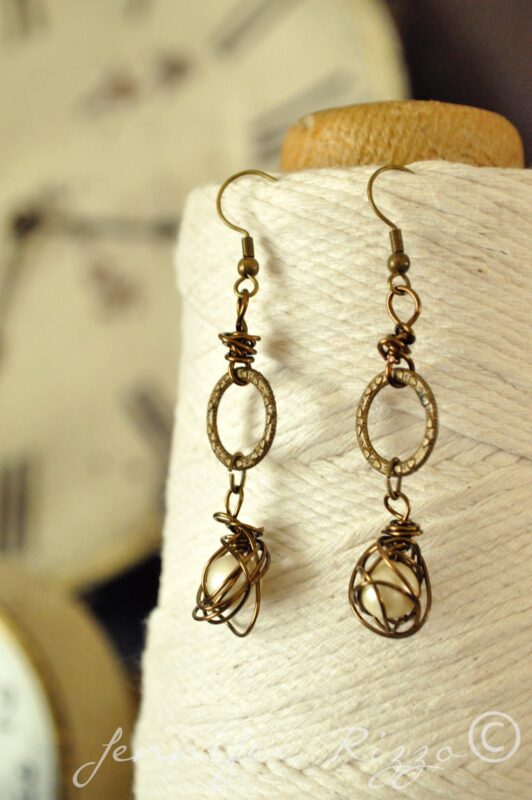 A pair of bronze wire-wrapped, handmade earrings with oval and bead details displayed hanging on a spool of cream-colored twine, inspired by Angel Santos Cage Dolls.