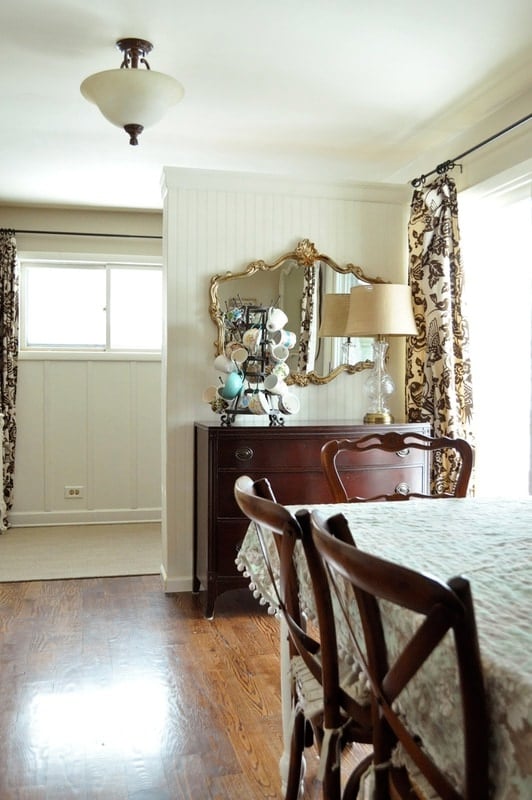 A dining room with wooden chairs and table, a sideboard with a lamp, decorative items, and a mirror on the wall, near a window with patterned curtains.