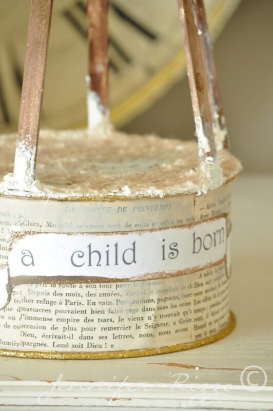 A cylindrical craft project decorated with vintage French newspaper text and the phrase “a child is born” in script of an art doll angel