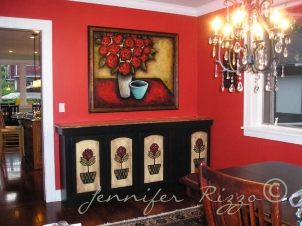 Dining room with red walls, a black sideboard featuring rose designs, a large floral painting, chandelier, and view into the kitchen. Painted wall murals add artistic flair. “Jennifer Rizzo” watermark across the image.