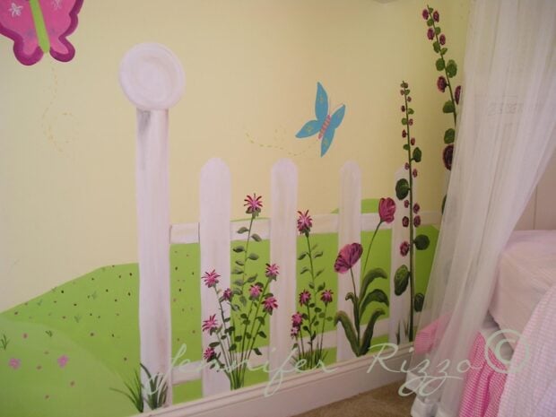 A painted wall mural of a white picket fence with flowers and butterflies decorates a light yellow bedroom wall next to a bed with a pink blanket.