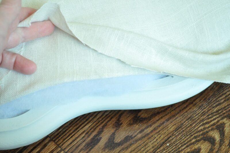 A hand lifts a piece of off-white fabric, revealing white padding and a curved base on a wooden floor—modernizing an old cane back chair with tufting.