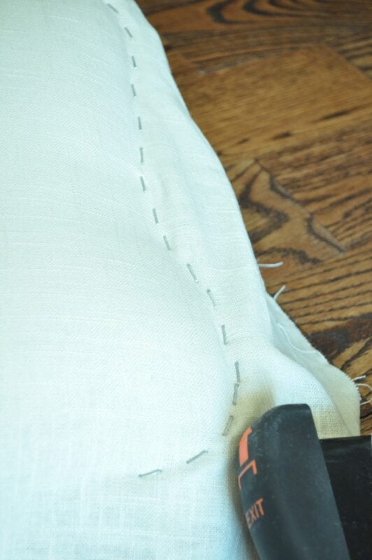 White fabric with visible hand-stitched basting along the edge, lying on a wooden surface next to the handle of a black tool—perfect for modernizing an old cane back chair with tufting.
