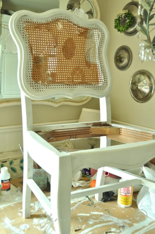 A white, partially painted wooden chair missing its seat sits on a cluttered surface with paint supplies and tools around it—perfect for modernizing an old cane back chair with tufting.
