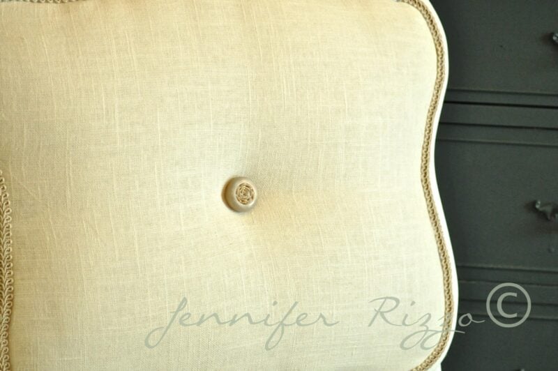 Close-up of a beige, square cushion with a single central button and decorative trim, perfect for modernizing an old cane back chair with tufting; placed in front of a dark piece of furniture.