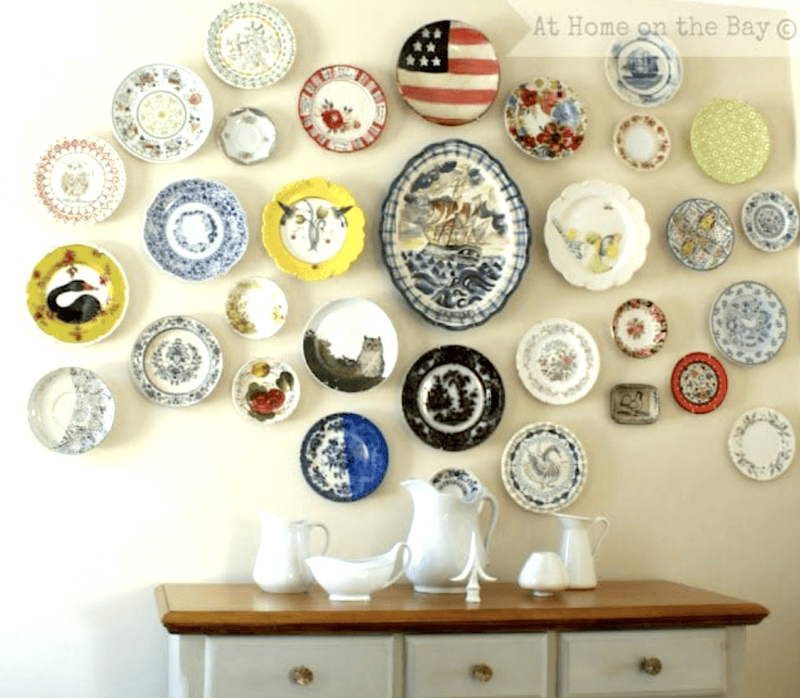 Assorted decorative plates create beautiful plate display walls, arranged in a pattern above a wooden sideboard topped with white ceramic pitchers and a bowl.