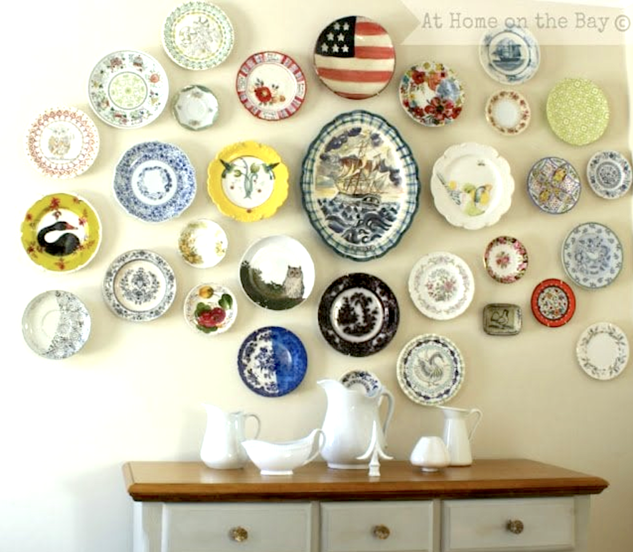 Assorted decorative plates create beautiful plate display walls, arranged in a pattern above a wooden sideboard topped with white ceramic pitchers and a bowl.