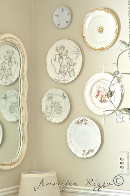 A group of decorative plates with floral and nature designs creates a beautiful plate display wall on a beige backdrop, accompanied by a mirror, chandelier, lamp, and electrical outlet.
