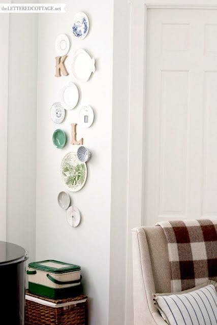 A beautiful plate display wall features assorted plates and wooden letters beside a closed door and an armchair draped with a plaid blanket. Nearby, green boxes are stacked on a basket for added charm.