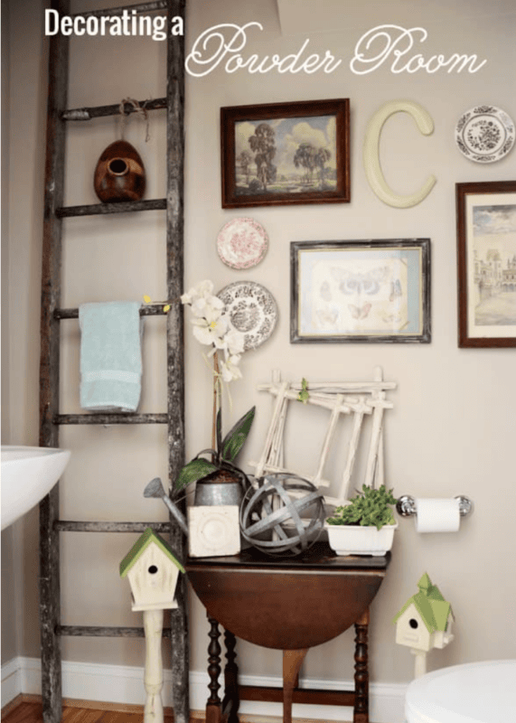 A decorated powder room features a rustic ladder with a towel, framed art, birdhouses, a small table, and a potted orchid beneath a “Decorating a Powder Room” sign—perfect inspiration for decorating a powder room with charm and style.