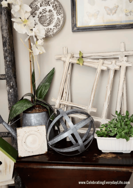 Decorative arrangement perfect for decorating a powder room: an orchid in a metal pot, a metal orb, empty distressed frames, a white planter with greenery, and vintage-style decor on a dark wooden table.