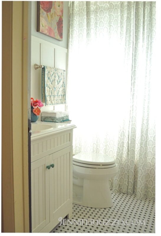 A bathroom from A Summer Tour of My Home with Flowers features a white cabinet, toilet, patterned shower curtain, black-and-white hexagonal tiles, and floral artwork alongside coordinating towels.
