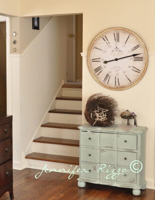 A large vintage-style wall clock hangs above a distressed blue dresser adorned with flowers, creating a warm welcome in the beige-walled entryway—just one stop on a summer tour of my home.