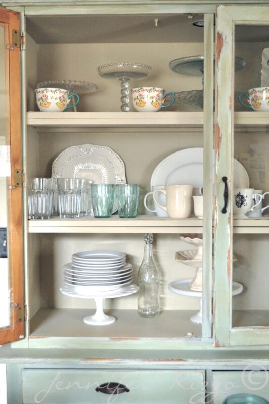 A vintage cabinet with glass doors displays stacked plates, cups, glasses, bowls, cake stands, and floral teacups on three shelves—a charming highlight from A Summer Tour of a Cottage home.