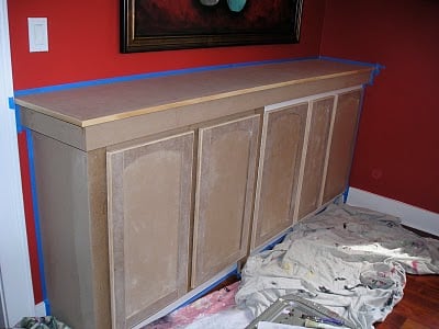 A partially painted wooden cabinet is taped off for painting in a room with red walls. Drop cloths, ideal for your hand-painted and stenciled furniture ideas, are spread on the floor below it.
