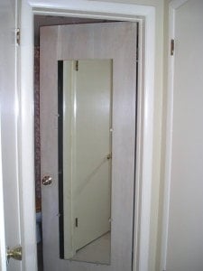 A partially open flat paneled door with trim and a full-length mirror attached reveals another closed door behind it in a white-walled room.