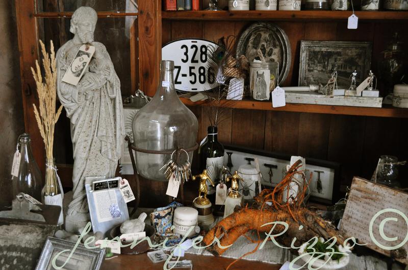 A shelf, inspired by the seven secrets to merchandising styling and display, showcases vintage items like a stone statue, glass jug, old license plates, bottles, dried plants, and assorted small antiques.
