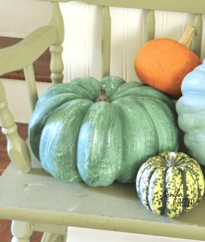 Three pumpkins—one large green, one small green-striped, and one orange—are arranged on a light green wooden chair in front of white paneling and wooden stairs, creating a charming scene perfect for painting fake pumpkins as autumn décor.