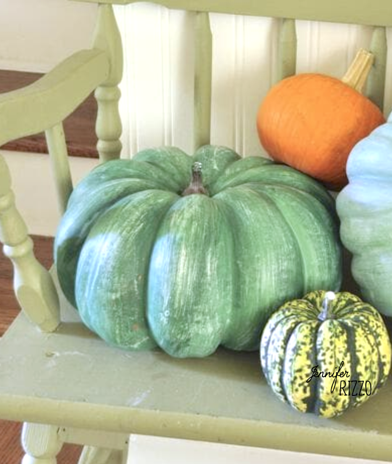 Three pumpkins—one large green, one small green-striped, and one orange—are arranged on a light green wooden chair in front of white paneling and wooden stairs, creating a charming scene perfect for painting fake pumpkins as autumn décor.