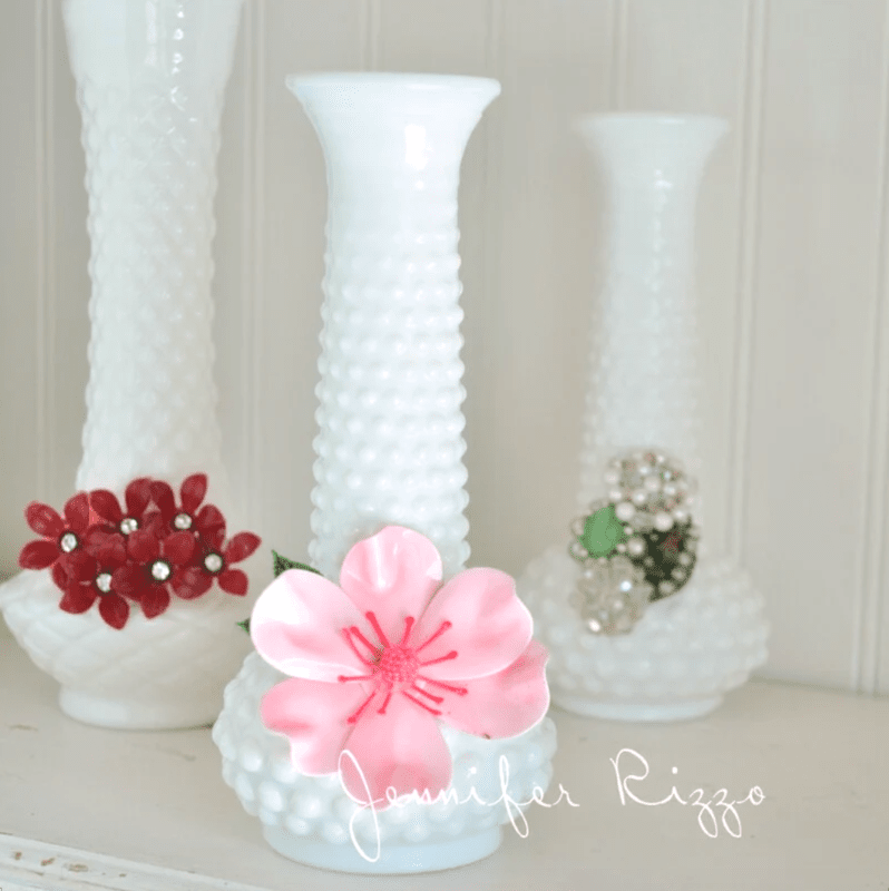 Three white hobnail-patterned vases are displayed, each upcycled from milk glass and adorned with a unique floral brooch made from broken jewelry.
