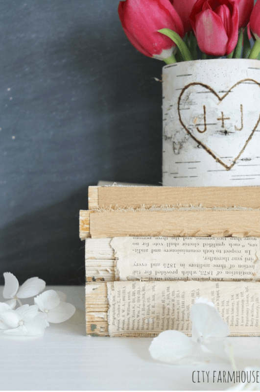 A rustic vase with red tulips and a carved heart, inspired by DIY birch bark vases, sits behind three stacked vintage books and scattered white petals on a light surface.