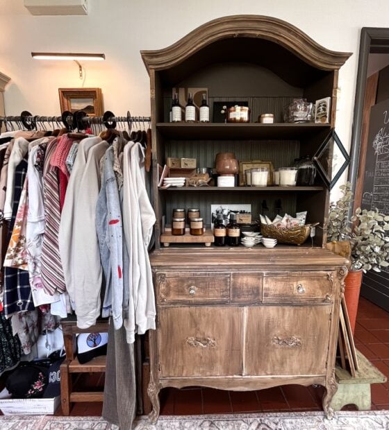 A wooden hutch, showcasing candles and jars, stands beside a clothing rack with shirts, sweaters, and scarves—offering stylish inspiration and repurposing ideas for outdated furniture in a chic boutique setting.