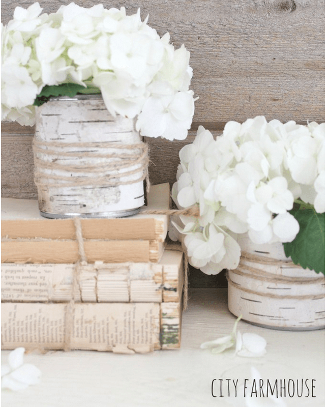 Two white hydrangea arrangements in DIY birch bark vases sit on and beside a stack of vintage books, against a rustic wooden background.