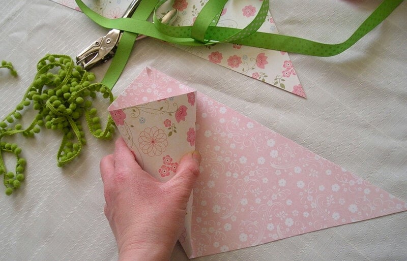 A hand demonstrates how to make a hanging paper cone vase, folding pink floral paper into a cone shape on a table with green ribbon, green yarn, and a hole punch nearby.