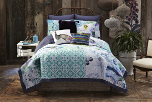 A bed adorned with Tracy Porter's signature patterned blue, green, and white bedding, accented by a purple pillow and a butterfly pillow. The room features wooden walls, vibrant plants, and eclectic decorative items.