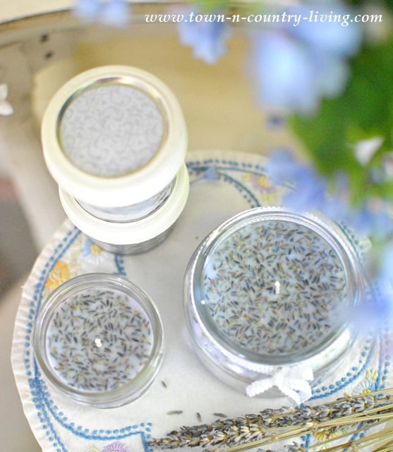 Three glass jars filled with a white substance and dried lavender buds stand elegantly on an embroidered cloth. Beside them lies a bundle of lavender and a blue-flowered plant, evoking the serene aroma of lavender scented candles.