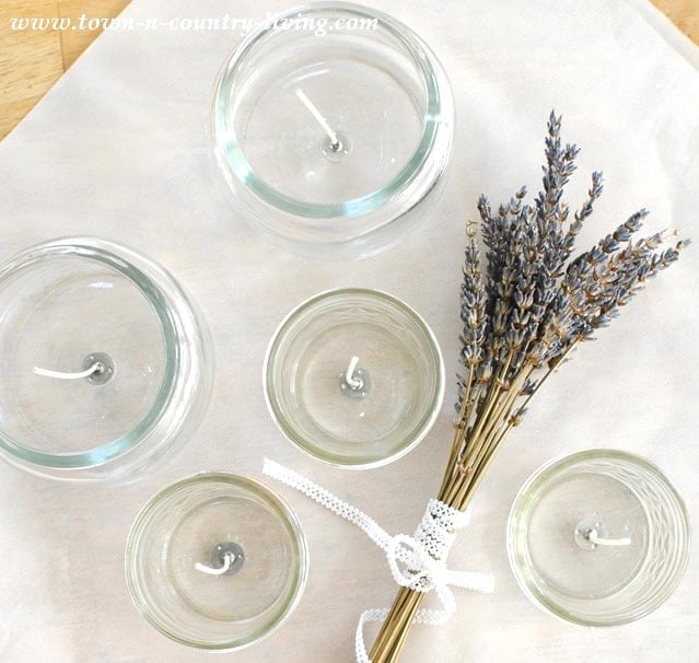 Five glass candle holders of varying sizes, each cradling lavender-scented candles, are arranged on a light cloth with a bundle of dried lavender tied with lace.
