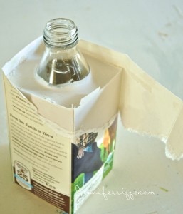 A clear glass bottle with a narrow neck is partially encased in torn cardboard packaging, subtly echoing the delicate charm of a vase made from plaster, all resting on a light surface.