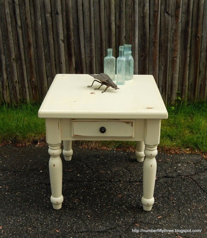 A small, vintage white wooden side table with a drawer, boasting a distressed makeover, stands on a pavement. On the table are three glass bottles and a decorative bird sculpture. A wooden fence and grass serve as the background.