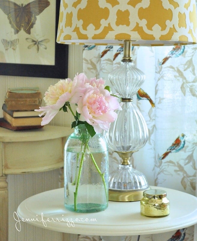 In this cozy home tour vignette, a glass jar with pink flowers graces a round table, complemented by a gold lamp and tiny gold jar. A stack of books and a framed butterfly print add charm in the background.