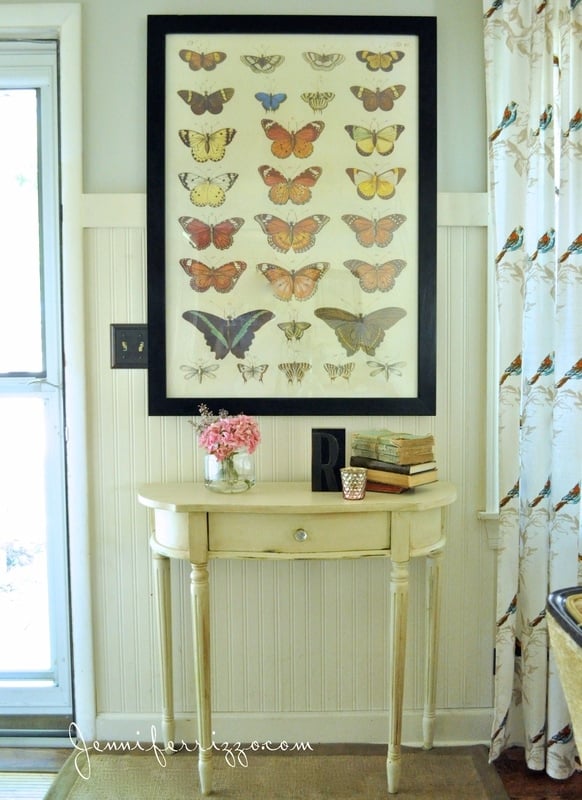 In this charming home tour, a wall adorned with framed butterfly art sits gracefully above a small cream-colored table, which holds a vase of pink flowers, books, and a decorative letter R. To the right, bird-patterned curtains complete the cozy vignette.