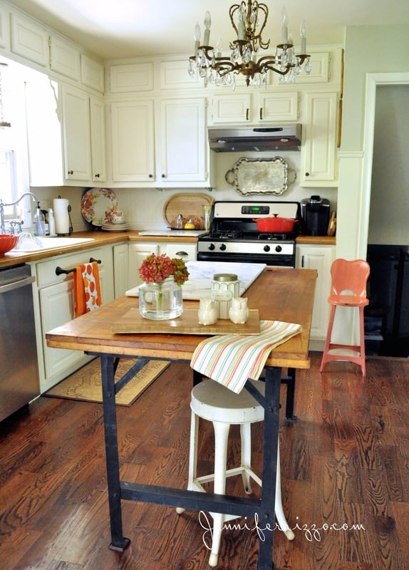 On this delightful home tour, a cozy kitchen showcases white cabinets and a wooden island with stools beneath an elegant chandelier. A pop of color comes from the red highchair amid the charming array of kitchen items.