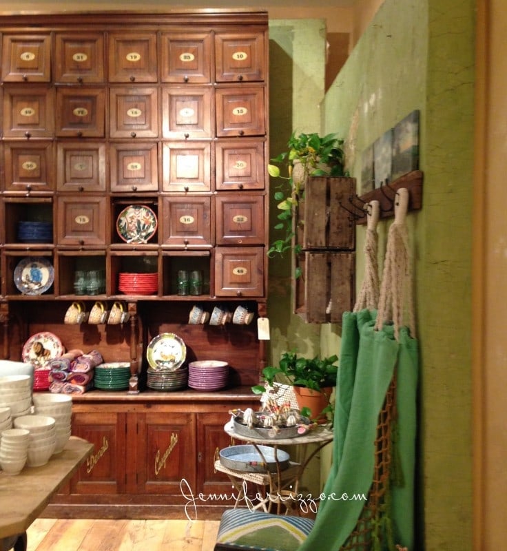 The cozy interior features a charming Anthropologie-style wooden cabinet filled with plates and cups, decorative dishes on display, and a small table adorned with potted plants. Green cloths hang gracefully on the wall, enhancing the inviting ambiance. Vintage Store Display Furniture
