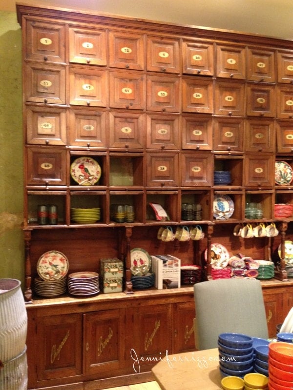 A charming Anthropologie-style wooden cabinet with multiple small drawers and shelves displays colorful plates, bowls, and decorative items.