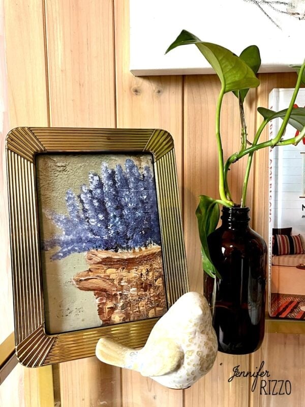 A gold-framed picture of purple flowers, a free art printable, a dark glass bottle with a green plant, and a ceramic bird ornament on a wooden shelf.
