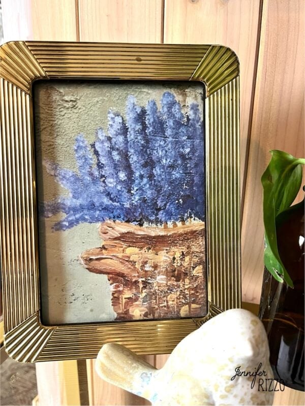 A gold-framed painting of purple-blue flowers in a brown vase sits on a wooden surface with a green plant and a ceramic bird nearby—available as a free art printable.