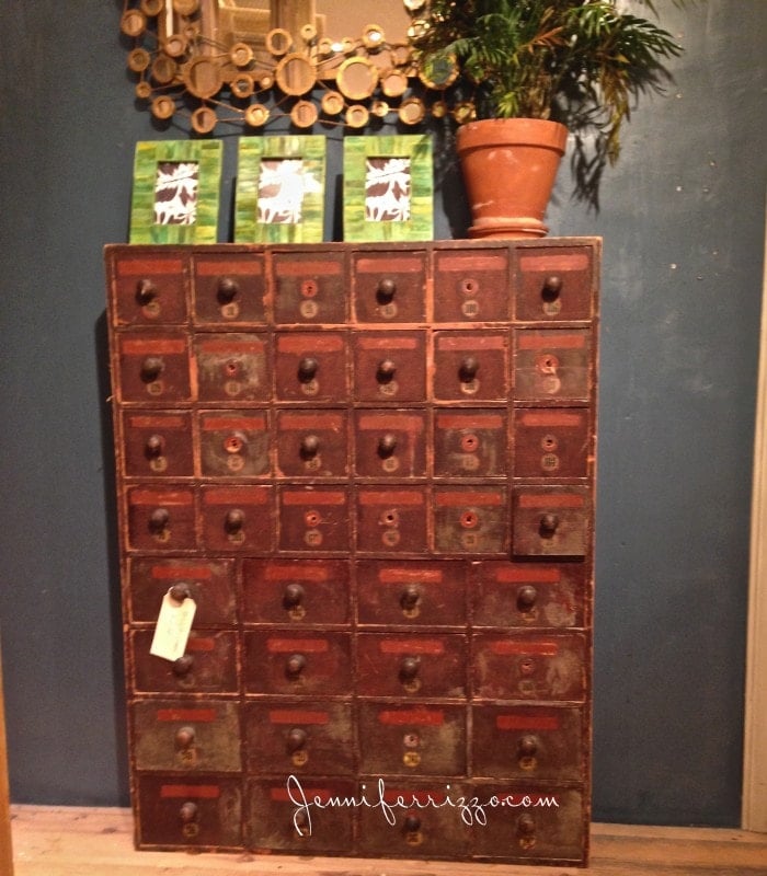 Vintage wooden apothecary cabinet with numerous small drawers, reminiscent of an Anthropologie find, a plant in a pot on top, and three framed images leaning against a wall.