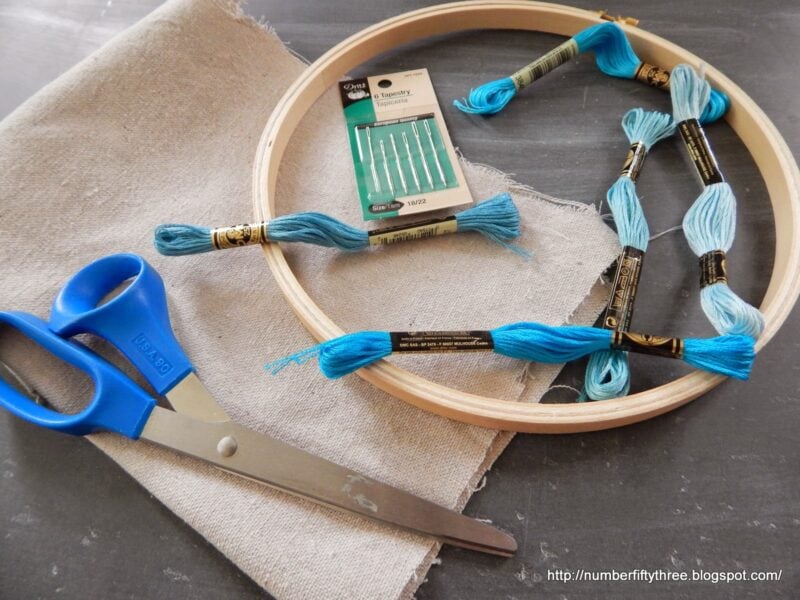 Easy DIY beginner embroidery project supplies including blue threads, a pack of needles, fabric, an embroidery hoop, and scissors lay neatly on a gray surface.