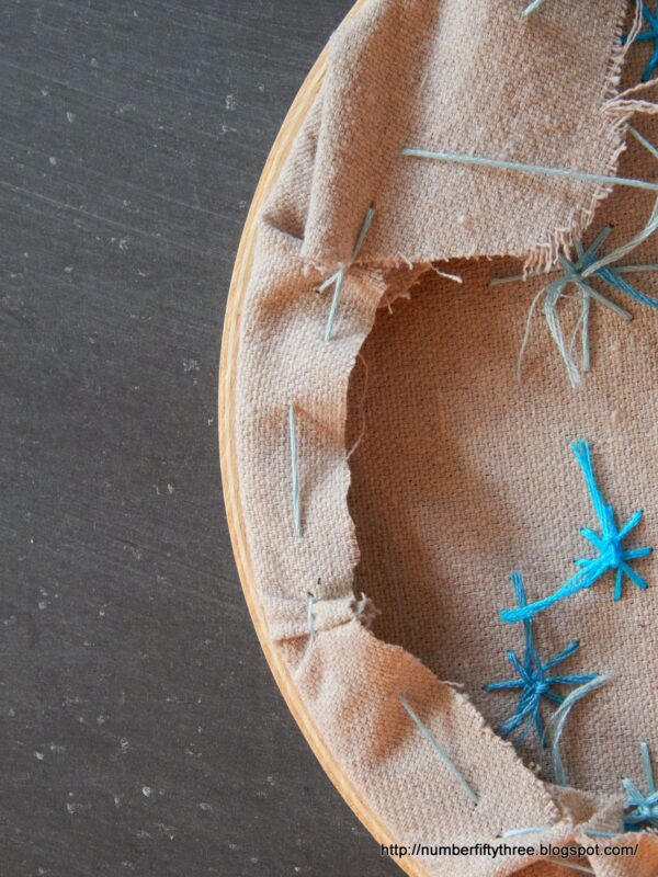 A close-up of tan fabric in an embroidery hoop, showcasing blue stitched patterns and loose threads—an easy DIY beginner embroidery project displayed on a dark surface.