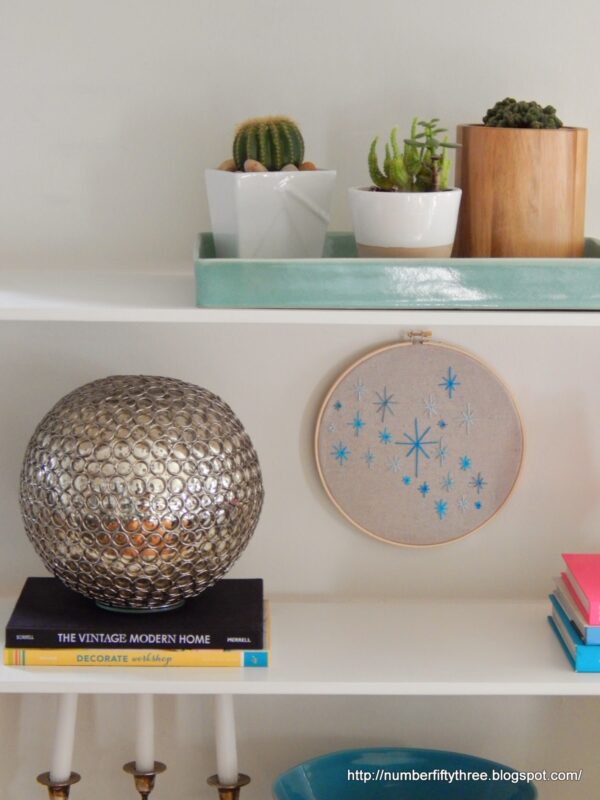 White shelves display stacked books, a silver decorative sphere, potted plants, a green tray, and an easy DIY beginner embroidery project—a stitched hoop adorned with blue stars.